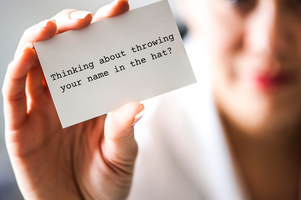 A person holding a card that says, "Thinking about throwing your name in the hat?"